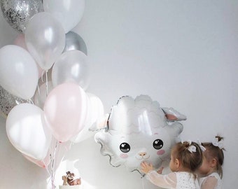 May include: Two young girls in white dresses with silver polka dots are standing in front of a table with a cake and a white sheep balloon. The balloon has a cute cartoon face with big eyes. There are silver and white balloons in the background.