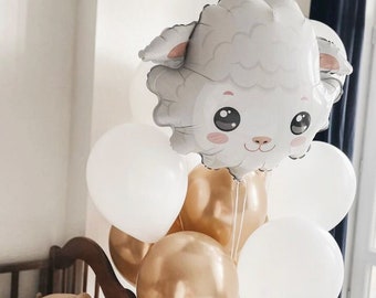 May include: A collection of balloons, including a large, gray sheep-shaped foil balloon with pink accents, and white and gold round balloons. The balloons are tied together with white string, creating a festive arrangement. A plush teddy bear is visible in the background.