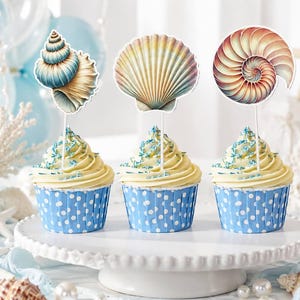 May include: Three cupcakes in blue and white polka dot liners topped with pale yellow frosting and blue sprinkles. Each cupcake features a decorative paper topper shaped like a seashell, including a spiral shell, a fan shell, and a nautilus shell.