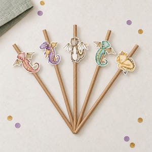 May include: Five brown paper straws arranged in a fan shape, each featuring a unique dragon cutout. The dragons are illustrated in red, purple, brown, green, and yellow, each with small wings and a tail, attached to the upper portion of the straws.