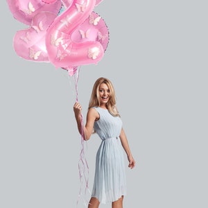 May include: A woman in a light blue dress holds pink balloons shaped like the number two, decorated with butterflies. The balloons are tied together with pink ribbons.