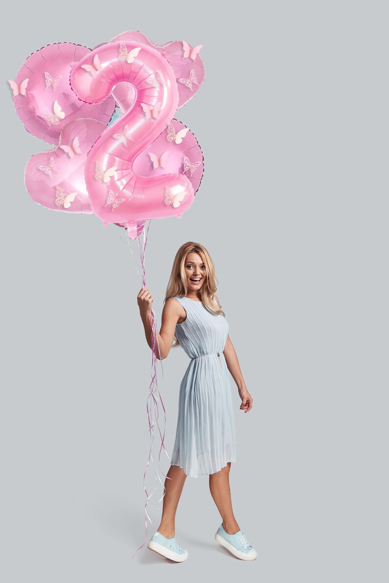 May include: A woman in a light blue dress holds pink balloons shaped like the number two, decorated with butterflies. The balloons are tied together with pink ribbons.