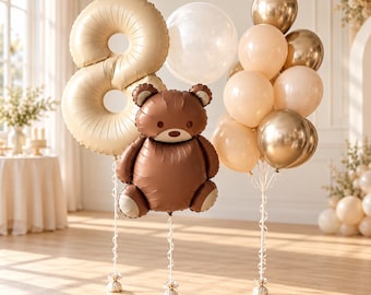 Puede incluir: Un arreglo de globos que incluye un globo de aluminio grande de color beige con el número ocho, un globo de aluminio con forma de oso de peluche marrón y un grupo de globos redondos en tonos crema, beige y oro metálico, atados con cintas blancas.