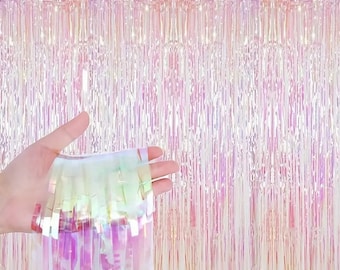 May include: A curtain of iridescent foil fringe in shades of pink, white, and purple. The fringe is hanging from a string and is being held in a hand.