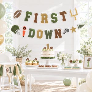 May include: A football-themed party banner with the text FIRST DOWN in green and gold letters, accompanied by football, helmet, whistle, and goalpost icons. A cake with a number one topper, cupcakes, and cookies are displayed on a table.