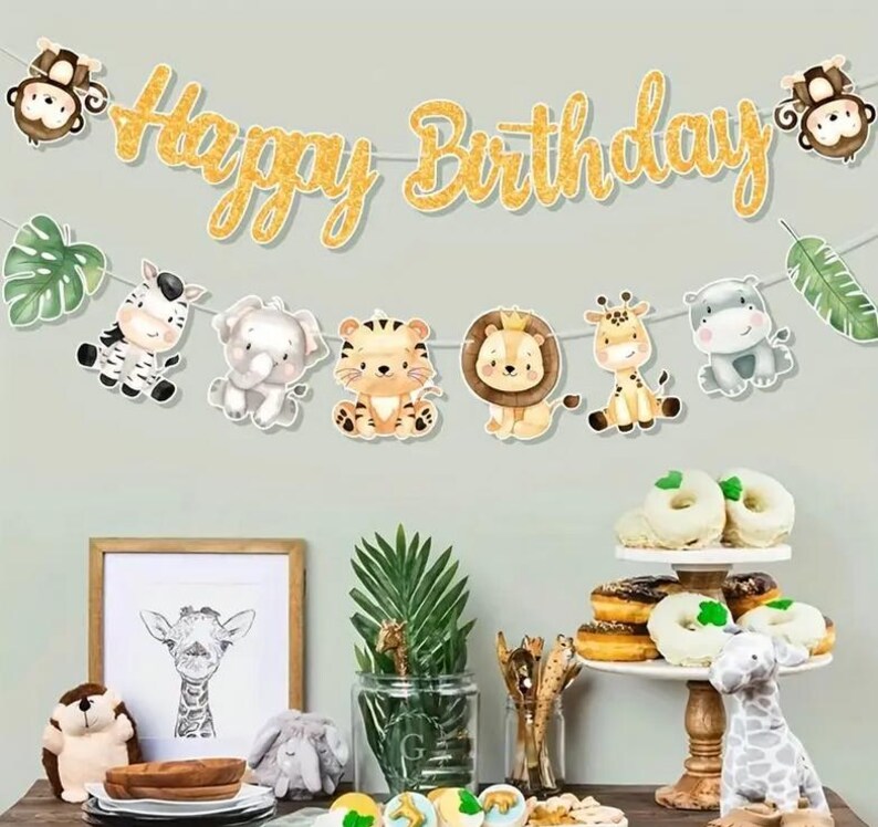 May include: A gold glitter "Happy Birthday" banner with cartoon animal decorations, including a zebra, elephant, tiger, lion, giraffe, and hippo. The image also features a framed giraffe print and a tiered dessert stand with doughnuts.