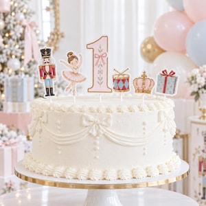May include: A white frosted cake topped with a set of decorative paper cutouts featuring a nutcracker, a ballerina, the number 1, a drum, a crown, and a gift box. The cake is decorated with piped white icing bows and swags around the sides.
