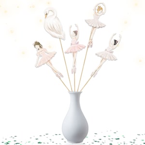 May include: A white vase holds several decorative ballet-themed toppers on wooden sticks. The toppers include a white swan and ballerinas in pink tutus. The ballerinas are in various poses, with their arms raised or extended. The background is white with gold stars.