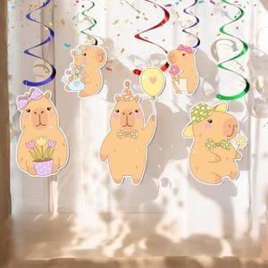 May include: A collection of cartoon capybara decorations. Each capybara has a unique design, including flowers, bows, hats, and bow ties. The decorations are in shades of tan, pink, and yellow, with colorful hanging streamers and confetti.