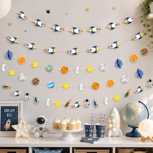 May include: A space-themed party decoration set featuring multiple strings of felt cutouts including rockets, planets, stars, moons, and astronauts. The set includes a framed sign with the text "DREAM BIG," a white astronaut figurine, cupcakes, two patterned paper cups, a rocket-shaped lamp, and a globe.