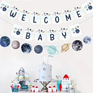 May include: A blue and white welcome baby banner with planets and stars. The banner is strung between two walls. There is a cake with a spaceman on top of it and other space-themed decorations on a table below.