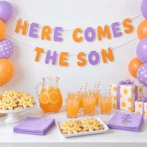 May include: A party banner with the text HERE COMES THE SON in orange and purple letters, surrounded by orange and purple balloons. The table features a pitcher and glasses of orange juice, sun-shaped cookies, and napkins with the text little brighter days ahead.