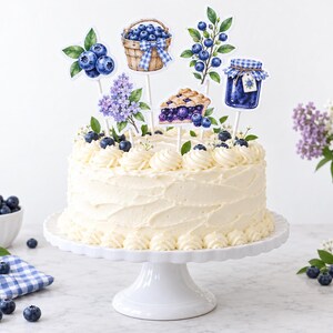 May include: A round white frosted cake on a pedestal stand, decorated with blueberry-themed paper toppers including a basket of berries, a slice of pie, a jar of jam, a sprig of blueberries, and a cluster of purple lilac flowers on sticks.