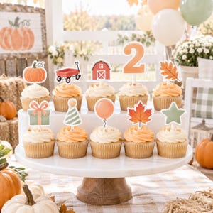May include: A set of ten cupcakes arranged on a white tiered stand, each topped with a decorative paper cutout featuring autumn themes including a pumpkin, red wagon, barn, number two, leaves, a gift, party hat, balloon, and a green star.