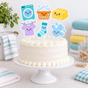 May include: A round white frosted cake on a white pedestal stand, topped with six decorative paper cutouts featuring laundry-themed illustrations including a washing machine, laundry basket, folded towels, a shirt, a sock, and soap bubbles.
