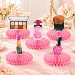 May include: A set of five pink honeycomb paper centerpieces featuring illustrations of a makeup palette, a perfume bottle with a red cap, a makeup brush, a lipstick tube, and a bottle of pink nail polish on a light pink surface.