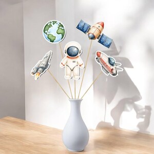 May include: A white vase holds space-themed cutouts on wooden sticks. The cutouts include an astronaut, a rocket, a satellite, and a planet Earth. The cutouts are in shades of white, blue, orange, and gray. The vase is on a wooden surface.