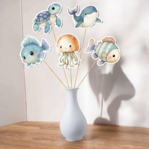 May include: A white vase holds wooden sticks topped with watercolor cutouts of sea creatures. The collection includes a turtle, whale, jellyfish, and two fish. The creatures are in shades of blue, orange, and beige, with smiling faces.