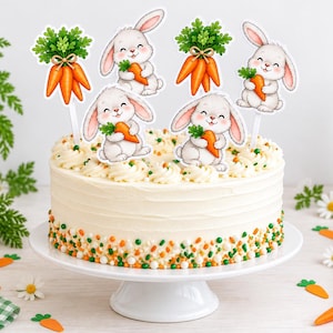 May include: A round cake on a white pedestal stand, decorated with white frosting and small orange and green sprinkles. Four paper cake toppers are inserted into the top, featuring illustrations of white bunnies holding orange carrots.