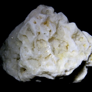 May include: A close-up of a light-colored, textured mineral specimen against a black background. The mineral has a rough, porous surface with small cavities and a slightly crystalline appearance. The color is primarily off-white with hints of yellow.