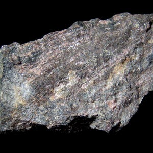 May include: A textured rock sample with a mix of dark grey, brown, and pink hues. The rock has a layered appearance with visible mineral grains and a small white label attached to the side.