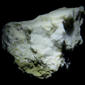 May include: A close-up of a white mineral specimen with a rough, textured surface. The mineral has areas of light green and yellow hues, set against a black background. The rock formation has a layered appearance.