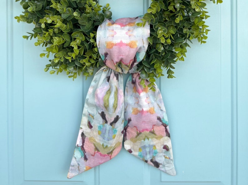 K&ouml;nnte beinhalten: Ein Kranz mit gr&uuml;nen Bl&auml;ttern und einer Schleife aus Stoff mit Blumenmuster. Die Schleife ist zu einem Knoten gebunden und die Enden h&auml;ngen herunter.