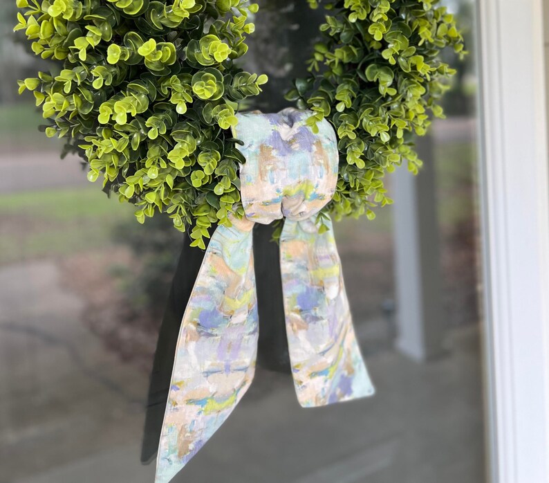 May include: A green wreath with a light blue, green, and beige patterned bow. The bow is tied in a knot and hangs down from the wreath.