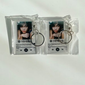 Op de afbeelding: Twee transparante acryl sleutelhangers met een zwart-witte afbeelding van een persoon en een Spotify muziekspeler ontwerp. Het muziekspeler ontwerp bevat de titel van het nummer "Slow Dancing (Layover)", de artiest "BTS-V" en een afspeelknop.