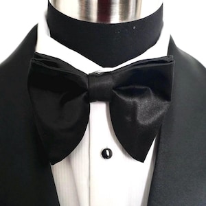 Könnte beinhalten: Ein schwarzer Satin-Fliege, Teil eines formellen Ensembles, steht im Mittelpunkt. Die Fliege ist ordentlich gebunden und sitzt über einem weißen Hemd, wobei eine schwarze Smokingjacke auf beiden Seiten sichtbar ist. Ein schwarzer Kragen und ein schwarzer Knopf sind ebenfalls zu sehen.
