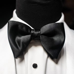 May include: A black satin bow tie is centered on a white dress shirt. The bow tie is knotted and has a slight sheen. The shirt has a black button and is on a black mannequin.