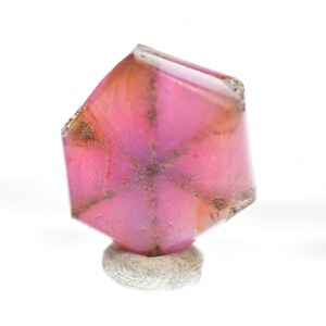 Puede incluir: Una piedra preciosa rosa en forma de estrella con un corte de seis lados y un patrón de estrella. La piedra preciosa está montada sobre una pequeña base gris texturizada.