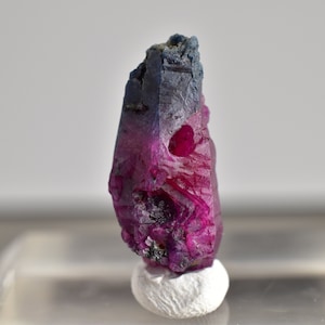 May include: A deep purple and pink crystal with a rough, faceted surface. The crystal is on a white stand.