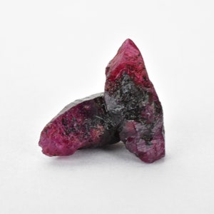 May include: Two raw ruby crystals, one larger than the other, with a deep red color and black inclusions. The crystals are on a white background.