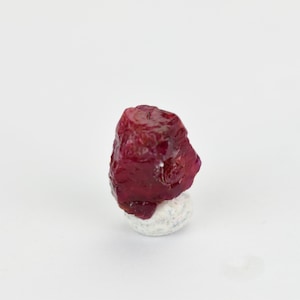 May include: A deep red ruby gemstone with a rough, natural finish. The ruby is displayed on a white pedestal.
