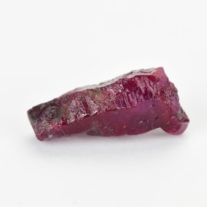 Può includere: Un cristallo di rubino rosso intenso con una texture ruvida e naturale. Il cristallo è traslucido e ha una leggera lucentezza.
