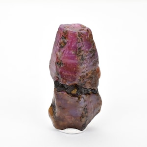 Pode incluir: Um cristal de rubi bruto, rosa e roxo, com uma inclusão marrom escura. O cristal tem aproximadamente 5 cm de altura e tem um topo pontiagudo.