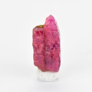 May include: A cluster of pink ruby crystals on a white base. The crystals are elongated and have a pointed shape.