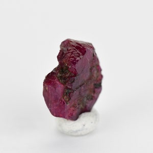 May include: A deep red ruby crystal with a rough, natural surface. The crystal is sitting on a small white pedestal.