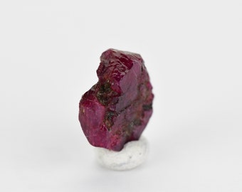 May include: A deep red ruby crystal with a rough, natural surface. The crystal is sitting on a small white pedestal.
