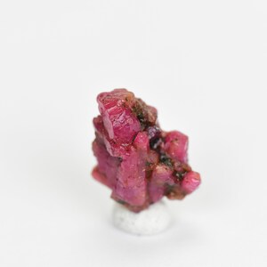 May include: A cluster of deep red ruby crystals on a white display stand. The crystals are faceted and have a glossy finish.