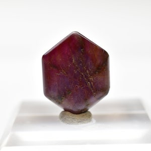 May include: A deep red, faceted gemstone with a hexagonal shape. The gemstone is displayed on a white stand.