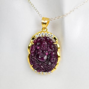 May include: A gold pendant necklace featuring a carved, deep red gemstone in the shape of a Buddha. The pendant is set in a gold frame with small, clear stones and hangs from a delicate silver chain.