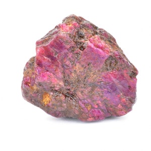 May include: A large, rough, pink ruby gemstone with a dark red hue. The ruby has a natural, unpolished appearance and is surrounded by a white background.