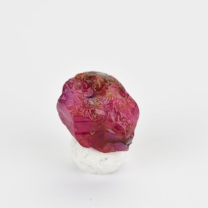 May include: A deep red ruby gemstone with a rough, faceted surface. The ruby is sitting on a white, round base.