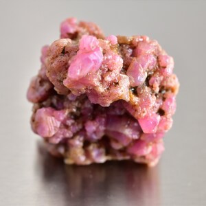 May include: A cluster of pink ruby crystals in a rough, natural state. The crystals are clustered together in a dense, irregular shape.