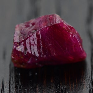 Puede incluir: Una piedra preciosa de rubí rojo intenso con una superficie rugosa y facetada. El rubí está sobre una superficie de madera oscura.