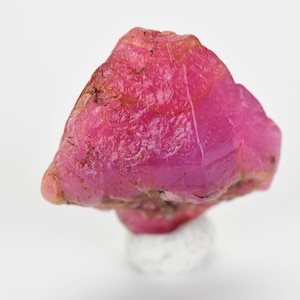 May include: A large, rough, pink ruby gemstone with a triangular shape. The ruby is on a white stand.
