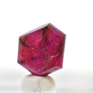 Puede incluir: Una gema facetada de color rosa intenso con forma hexagonal. La gema está apoyada sobre una superficie blanca.