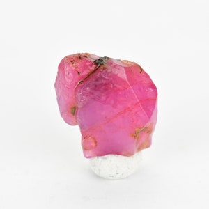 May include: A pink ruby crystal with a rough, natural appearance. The crystal is on a white stand.
